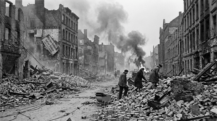 Three people walking over rubble in heavily damaged city street with smoking ruins