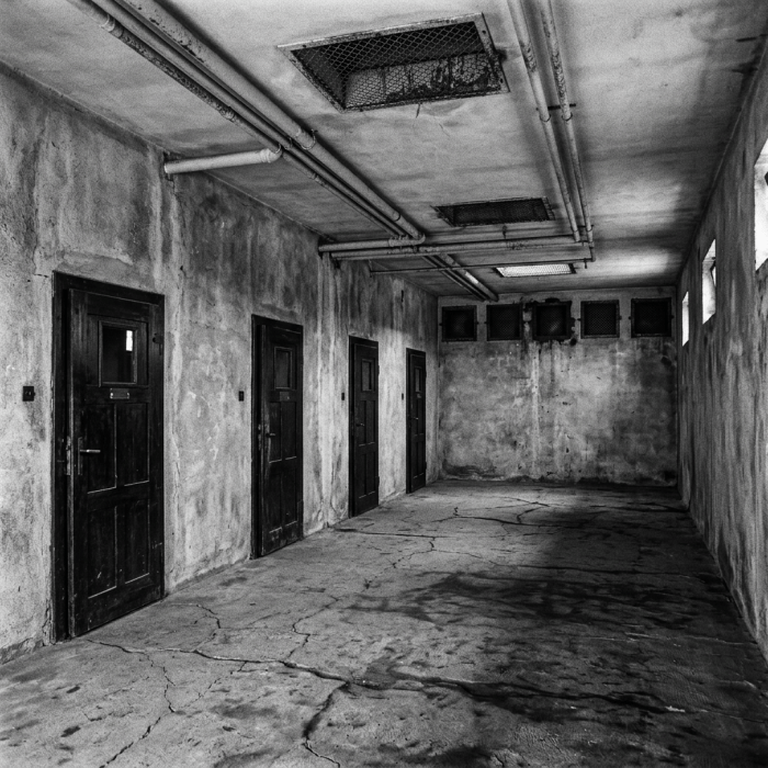 Long narrow corridor with four closed wooden prison cell doors on one side and small barred windows near the ceiling on the other side