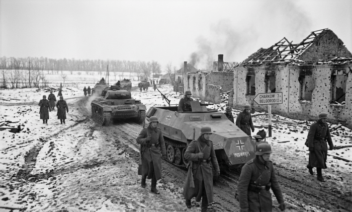 German soldiers and armored vehicles pass snowy ruins; sign reads CHARKOW 3 m, BELGOROD 12 m.